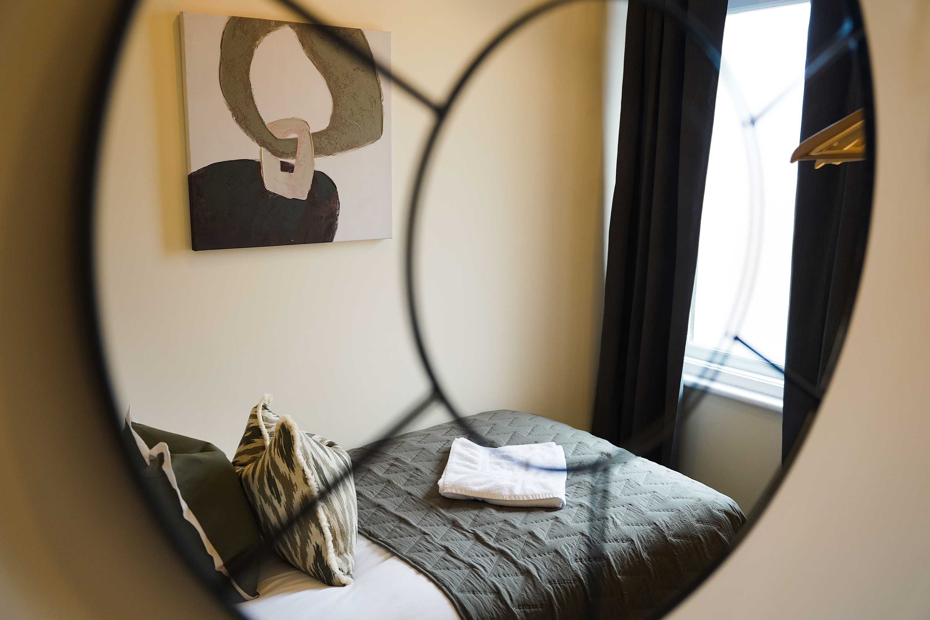 Una habitación con cama, almohada, manta, un cuadro en la pared y una ventana con cortinas.