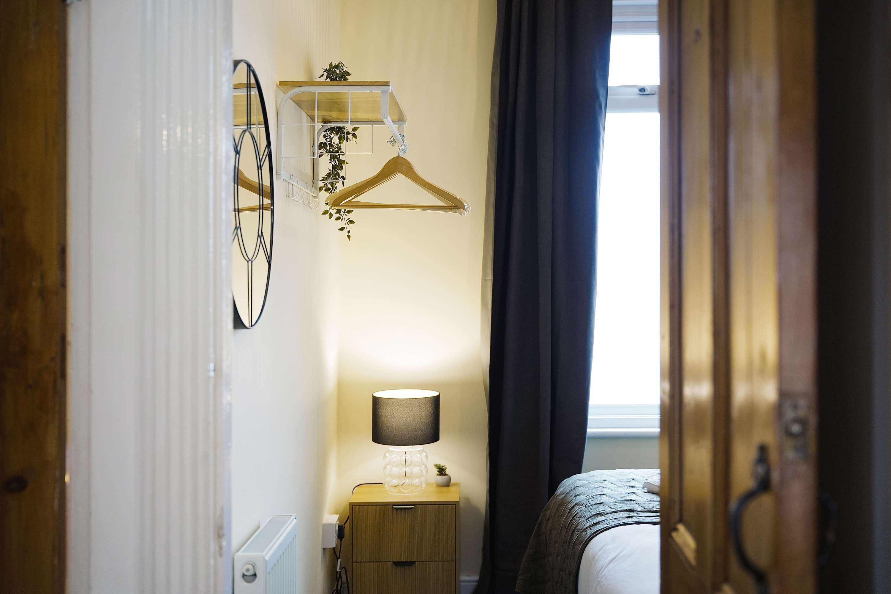 Een hotelkamer met een bed, een nachtkastje met een lamp, een spiegel aan de muur en een houten kapstok.