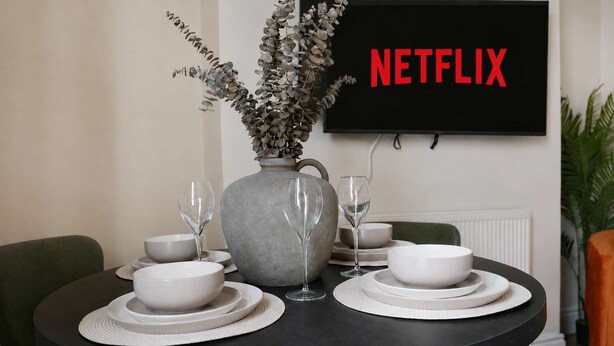 A dining table with white plates, bowls, and glasses, a grey vase with dried flowers, and a black wall-mounted TV displaying 'NETFLIX'. - Elegant 4BR Houghton St Sleeps 7 by Stay With Us (Hartlepool)