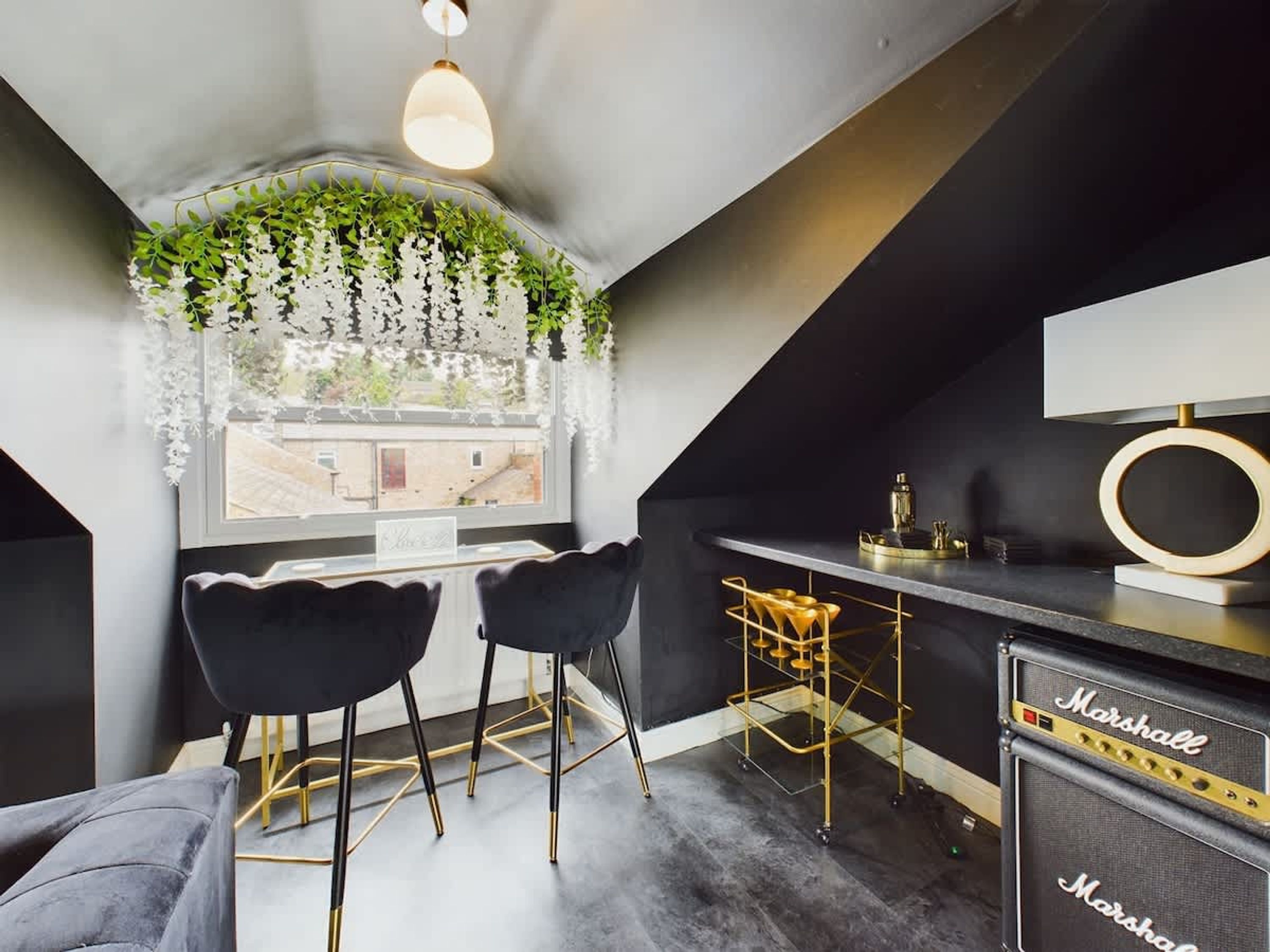 Une cuisine moderne aux couleurs noir et or, avec deux tabourets de bar noirs, une plante suspendue et une vue par la fenêtre.