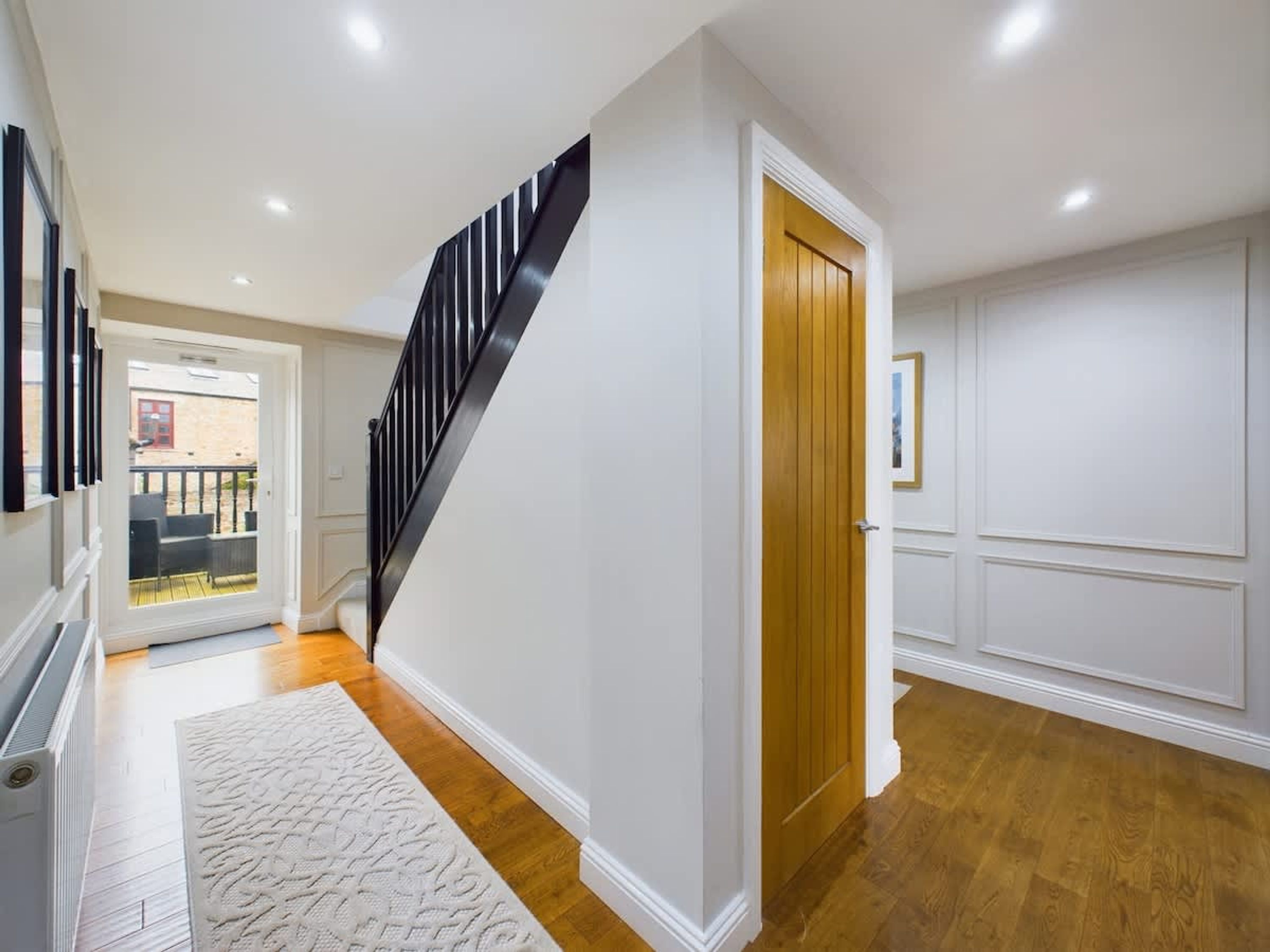 A hallway with a wooden floor, a white rug, a black staircase, and a wooden door.