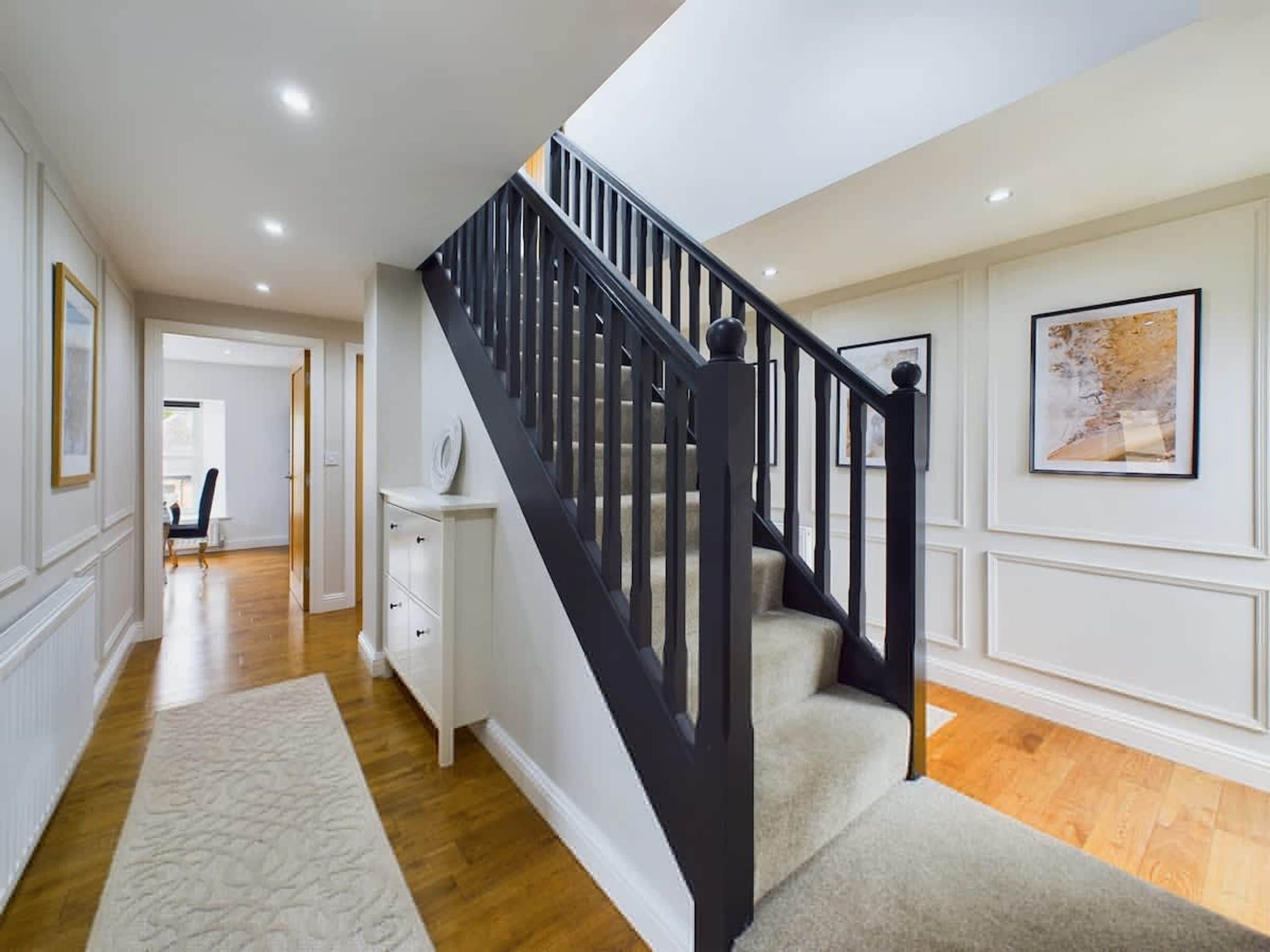 Un escalier avec des rampes noires menant à un deuxième étage, un mur blanc orné d’un tableau encadré et un plancher en bois.