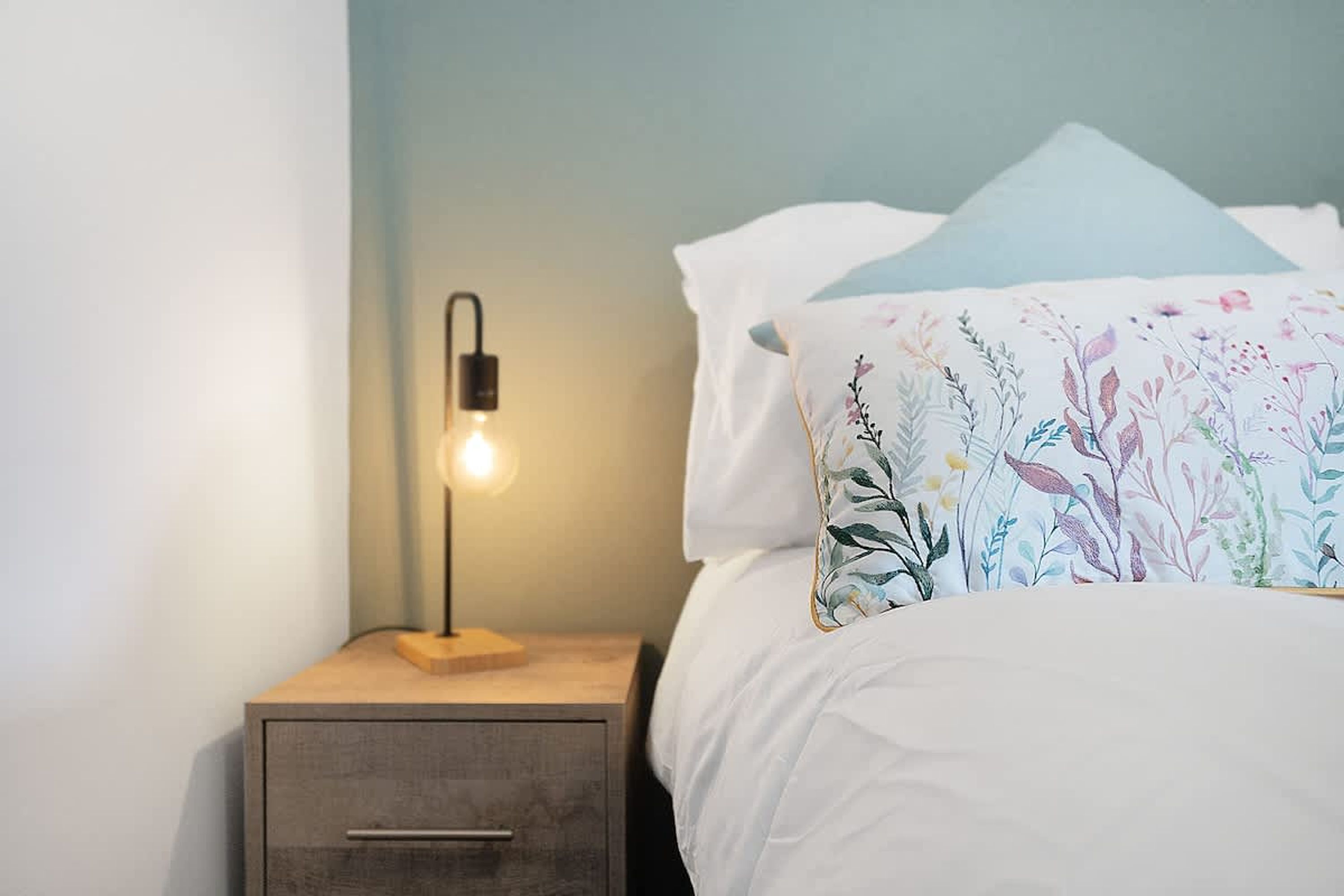 A neatly made bed with a floral pillow, a bedside table with a lamp, and a drawer.