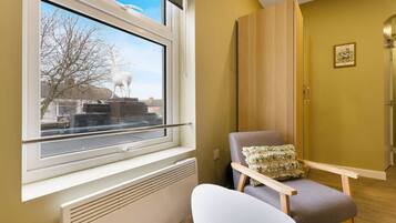 A room with a window, a radiator, a chair with a cushion, a wooden wardrobe, and a framed picture on the wall.
