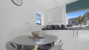 Een moderne keuken met witte kastjes, een ronde eettafel met stoelen, een klok aan de muur en uitzicht op de stad door een raam.