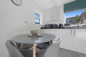 A modern kitchen with white cabinets, a round dining table with chairs, a clock on the wall, and a view of the city through a window. - Stylish Home Near Durham Heritage Coast Sleeps 5 (Durham)