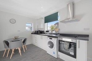 A compact kitchen with a washing machine, oven, and a dining area with a table and chairs. - Stylish Home Near Durham Heritage Coast Sleeps 5 (Durham)