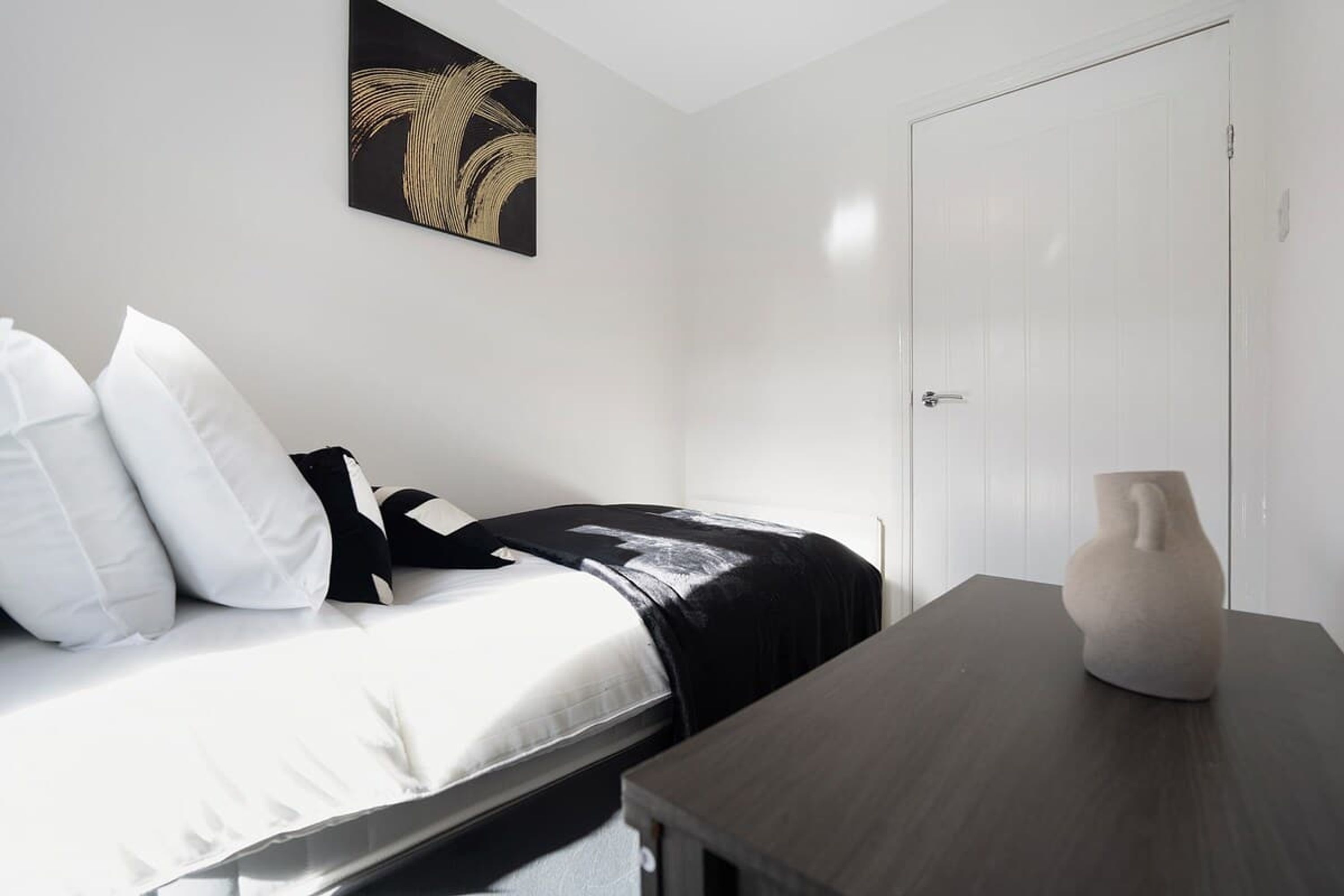 A bedroom with a bed, pillows, a bedside table, a vase, and a door.