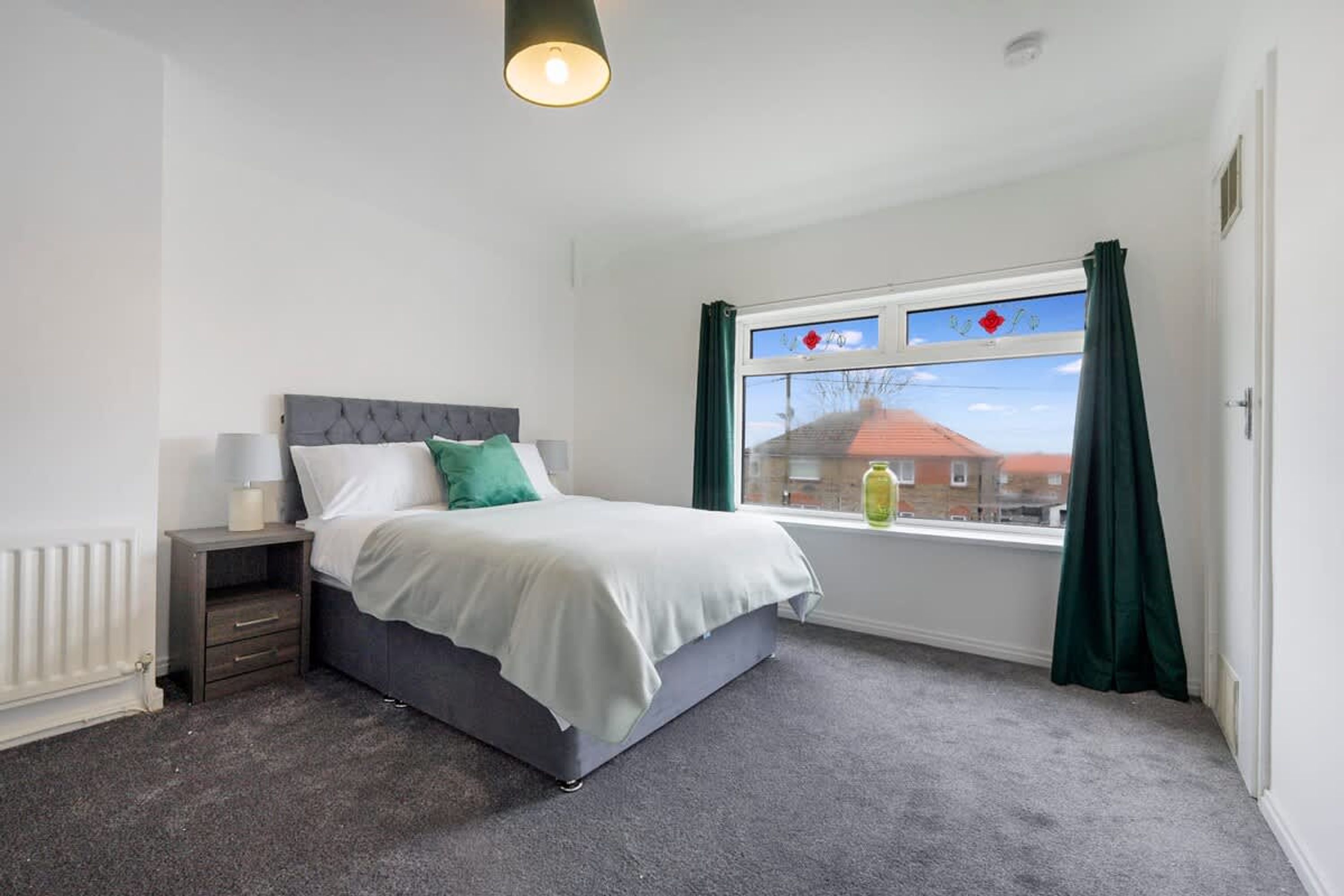 A bedroom with a bed, a nightstand, a window with curtains, and a radiator.