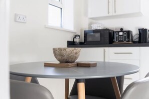 A modern kitchen with white cabinets, a microwave, a coffee maker, and a round table with a bowl and coasters. - Stylish Home Near Durham Heritage Coast Sleeps 5 (Durham)