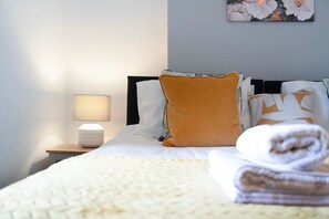 Ein ordentlich bezogenes Bett mit weißer Decke, einem gefalteten Handtuch und einer Nachttischlampe auf einem Nachttisch.