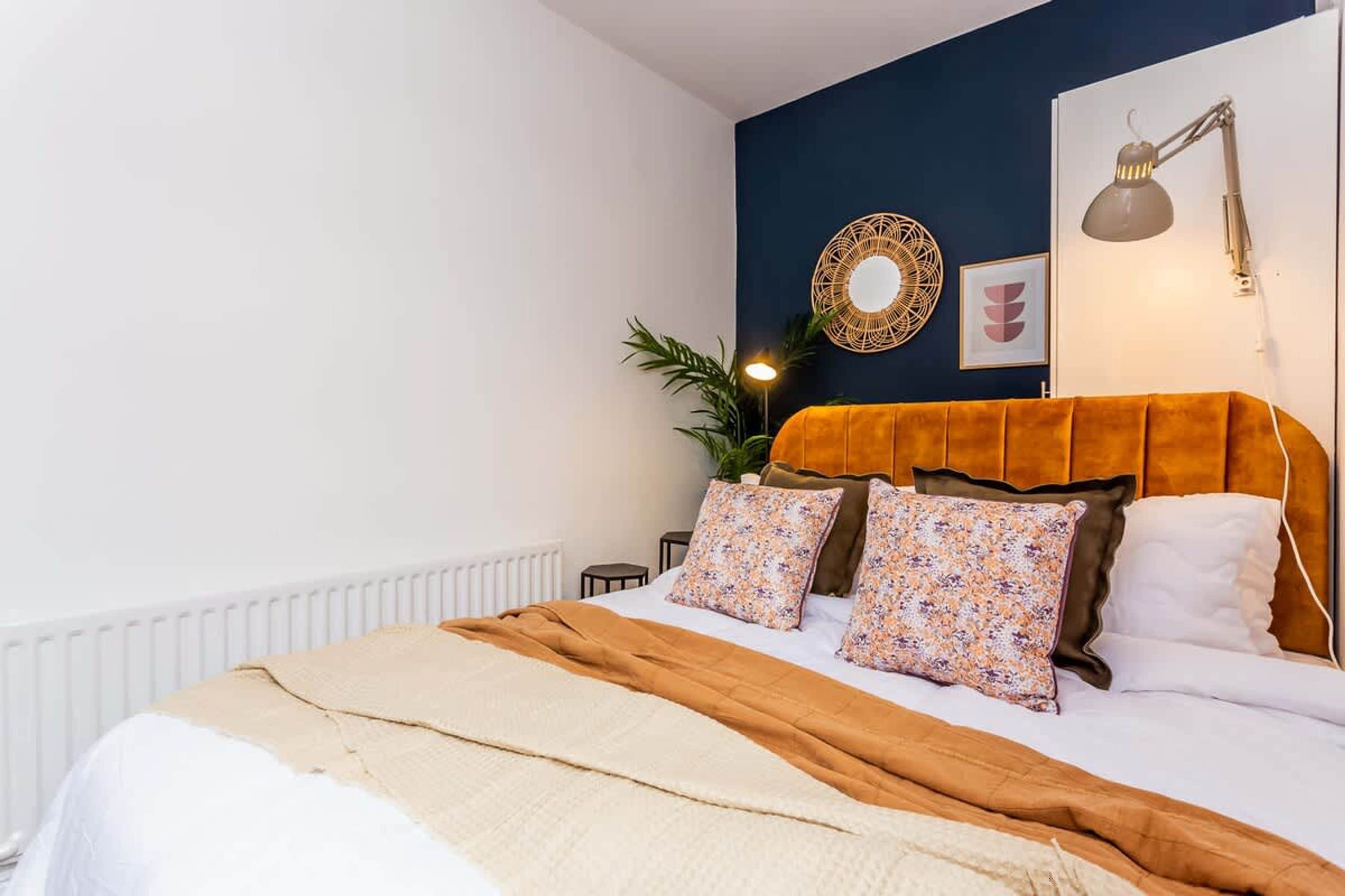 Un dormitorio con cama, lámpara de noche, espejo en la pared y una planta en maceta.