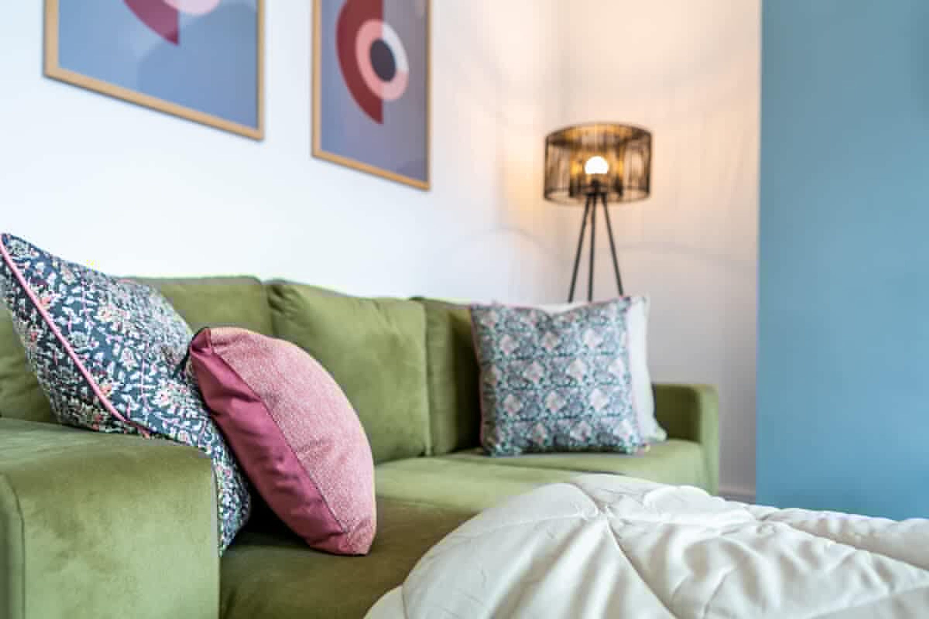 A green sofa with patterned cushions, a floor lamp, and a bed with a white cover.