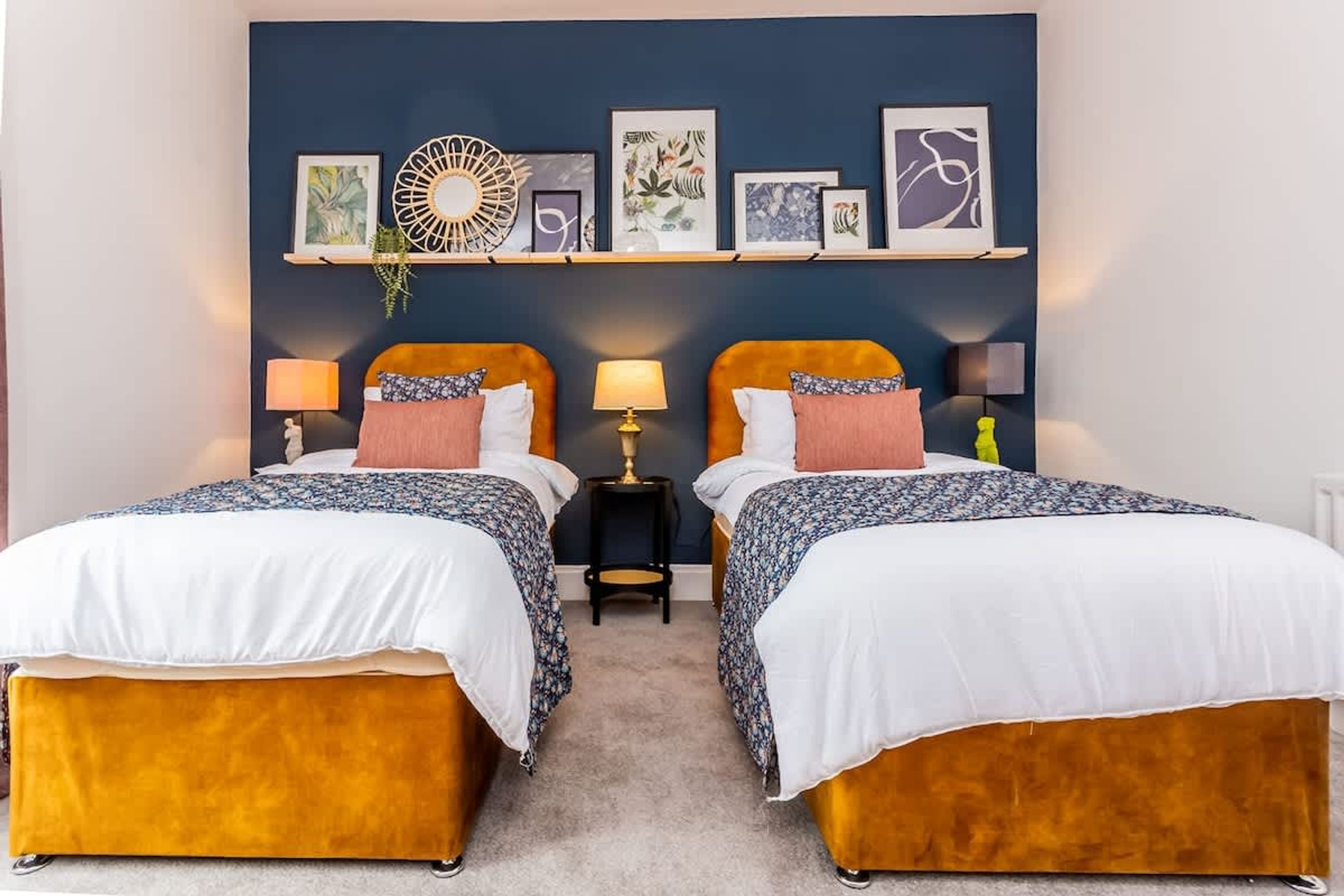 Habitació d'hotel amb dos llits, una petita taula de nit amb una làmpada, i una paret decorada amb obres d'art enmarcades.