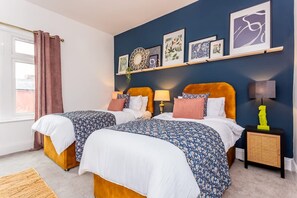 A double room with two beds, a window with curtains, a wall with artwork, and a small table with a lamp. - Two Bedroom Family Apartment Near Town Centre Chic Stay (Darlington)