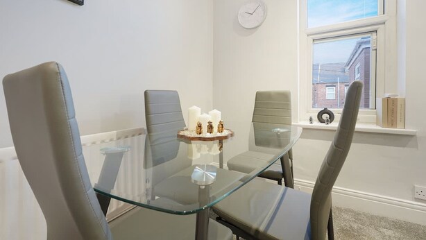 A dining area with a glass table, four chairs, a clock on the wall, and a window with blinds. - Chic 2 Bed With Wifi Parking Ideal for Work Stay (South Shields)