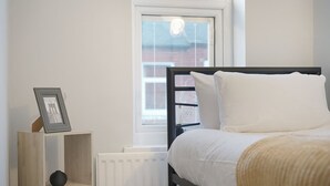 A bedroom with a bed, a bedside table with a picture frame, a radiator, and a window with blinds. - Chic 2 Bed With Wifi Parking Ideal for Work Stay (South Shields)