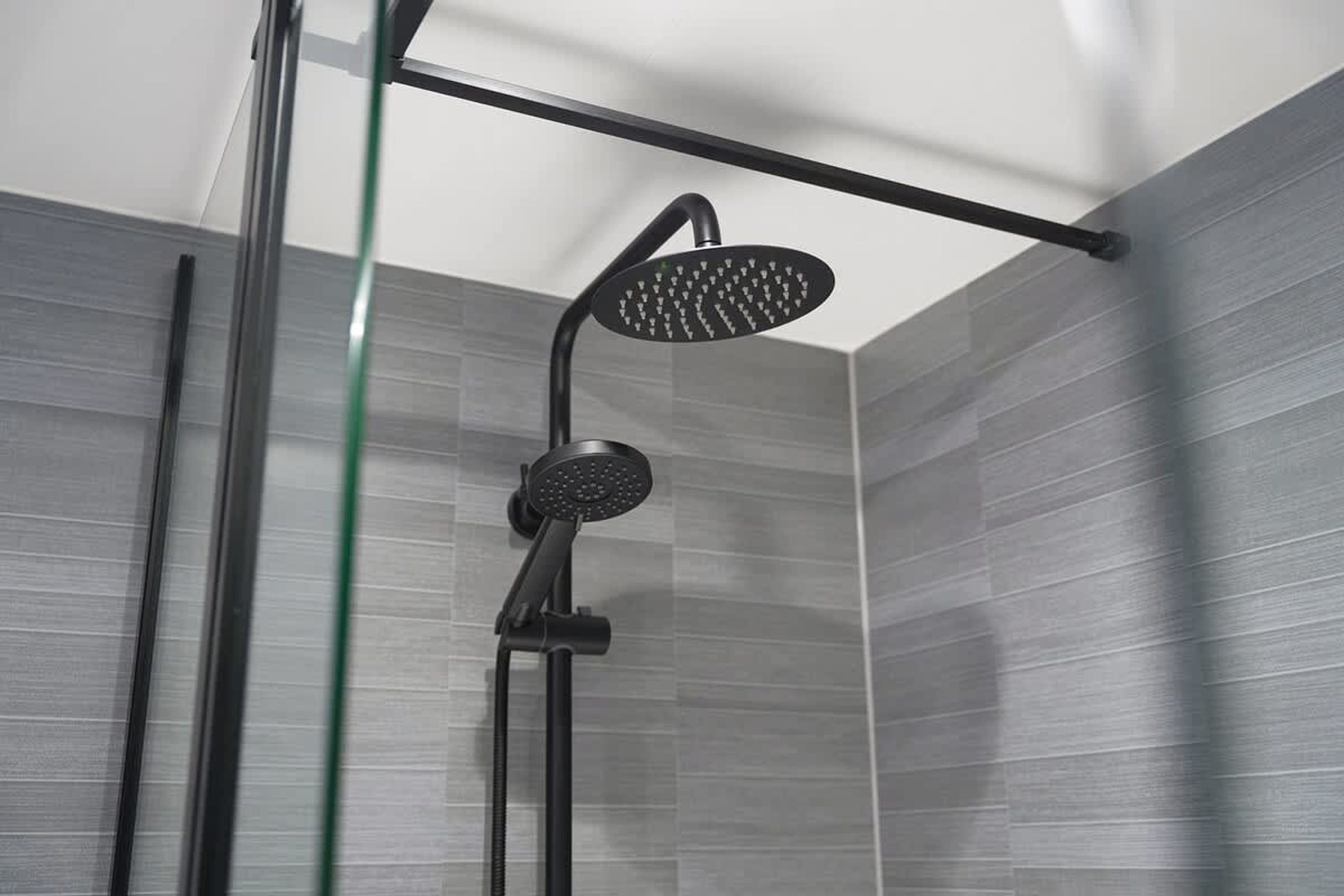 Une douche moderne dotée d’un pommeau de douche à main et d’une douchette à pluie fixée au plafond, le tout installé sur un mur carrelé.