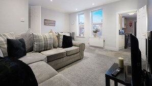 A living room with a sofa, a remote control on a table, and a window with a view of the outside. - Chic 2 Bed With Wifi Parking Ideal for Work Stay (South Shields)