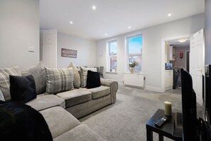 A living room with a sofa, a remote control on a table, and a window with a view of the outside. - Chic 2 Bed With Wifi Parking Ideal for Work Stay (South Shields)