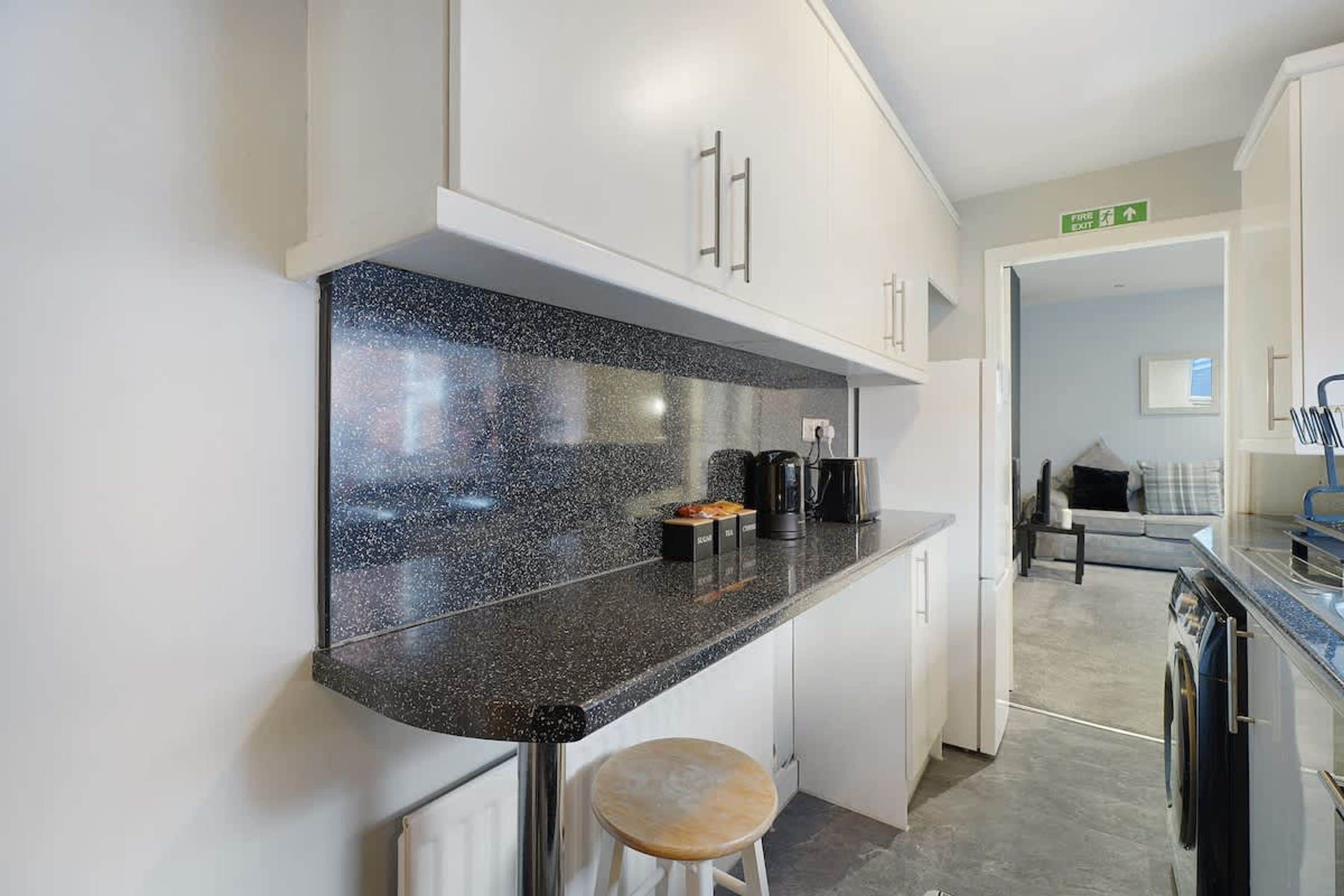 A modern kitchen with a granite countertop, stainless steel appliances, and a wooden stool.