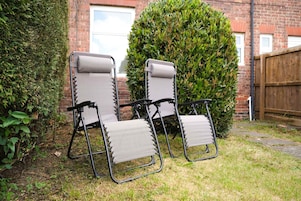Two outdoor lounge chairs with a metal frame and fabric seat and backrest, positioned on a grassy lawn with a brick wall and a wooden fence in the background.