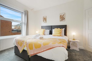 A hotel room with a bed, two bedside lamps, a window with curtains, and two wall-mounted artworks.
