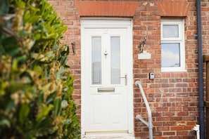 A white door with a number plate, a small window with a welcome sign, and a metal pipe on the brick wall. - Modern 3BR Washington Sleeps 6 by Stay With Us (Washington)
