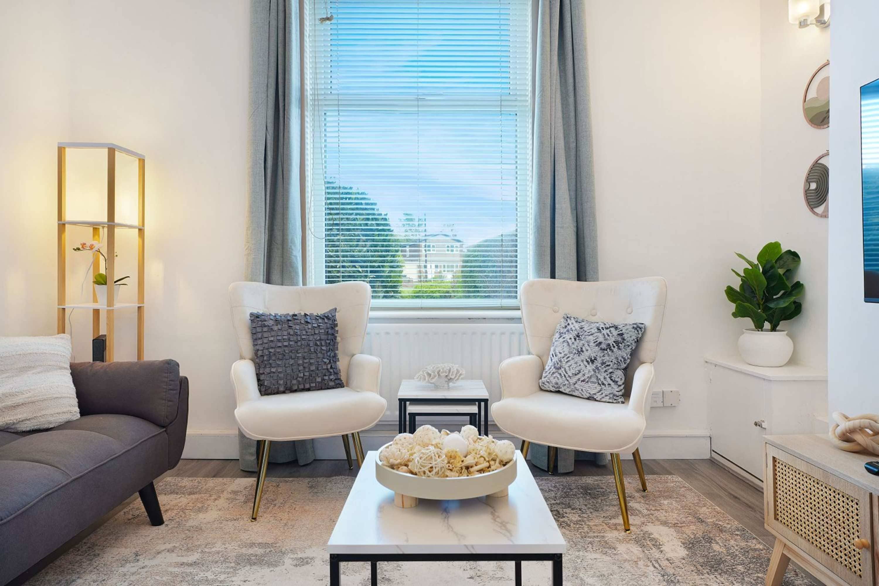 Un salon moderne avec deux fauteuils blancs, un canapé gris, une table basse en marbre et une vue sur les immeubles par la fenêtre.