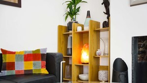 Uma sala de estar com um sofá preto, uma almofada colorida, uma mesa de centro de vidro e uma estante de madeira com diversos objetos decorativos.