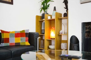 A living room with a black sofa, a colorful cushion, a glass coffee table, and a wooden shelving unit with various decorative items. - Chic 3BR Rotherham Parking Wifi By Stay With us (Rotherham)