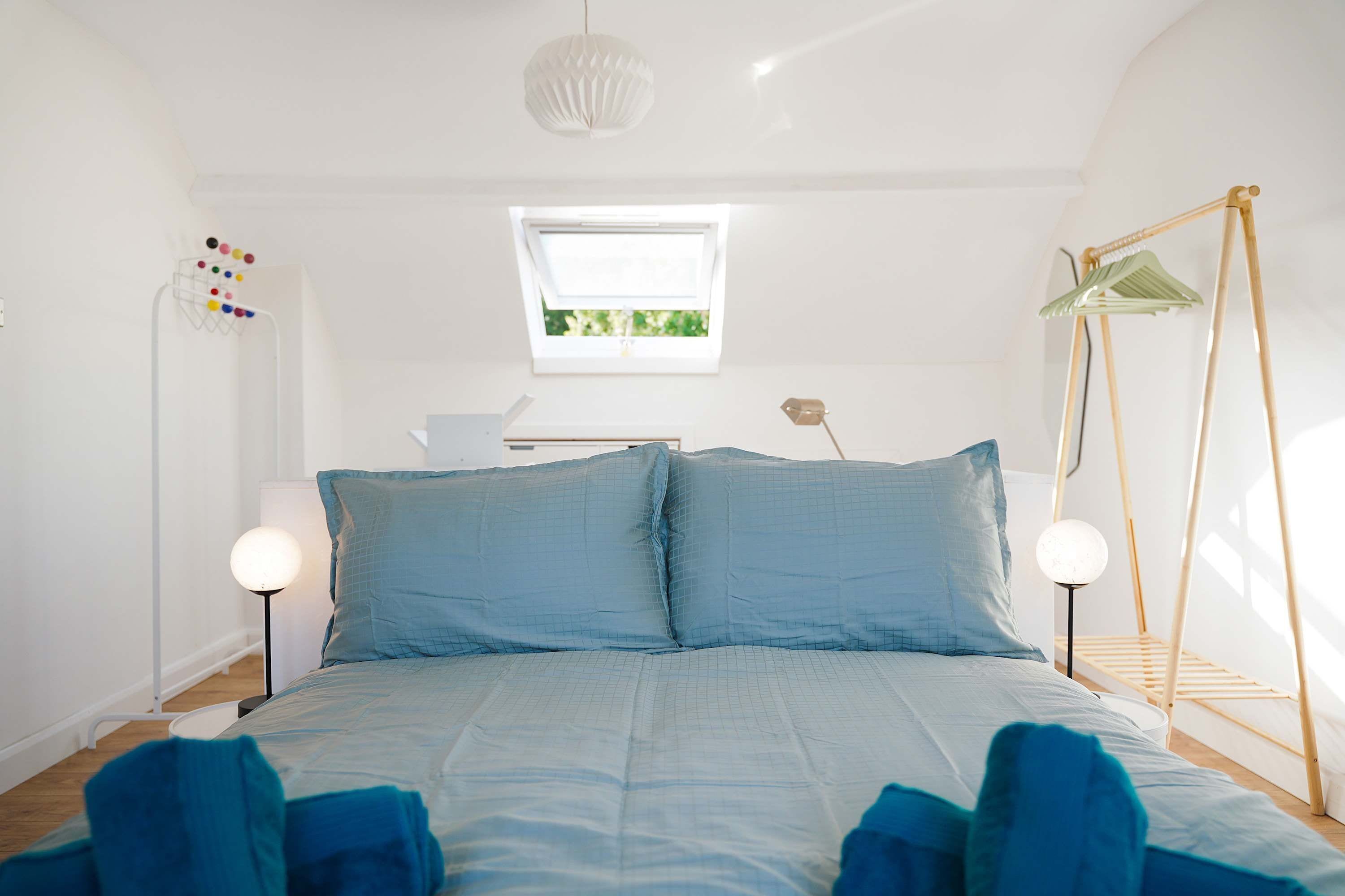 A bedroom with a bed, bedside lamps, a hanging light fixture, a window, and a clothes drying rack.