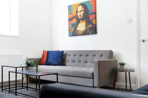 Una sala de estar con un sofá gris, dos mesitas auxiliares, una mesa redonda pequeña y un cuadro de una obra de arte famosa en la pared.