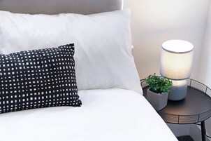 Una cama con ropa blanca y una almohada negra con diseños blancos, una mesita de noche con una lámpara y una planta en maceta.