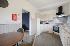 A compact kitchen with white cabinets, a microwave, a stove, and a refrigerator. There is a dining table with chairs in the foreground. - Work Friendly 3 Bed House With Sofa Beds Sleeps 7 (Stockton-on-Tees)
