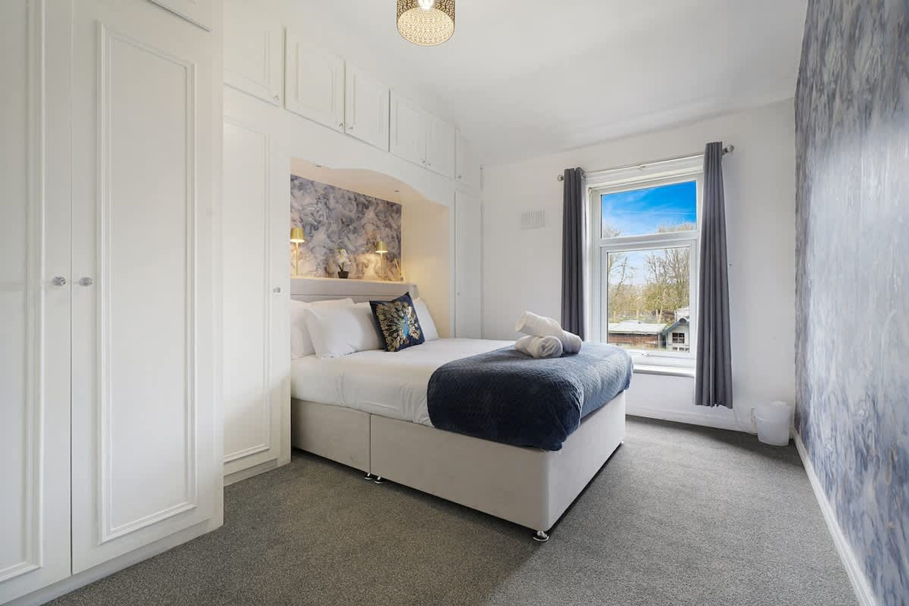 A bedroom with a bed, a window with curtains, a wall-mounted shelf with lamps, and a door.