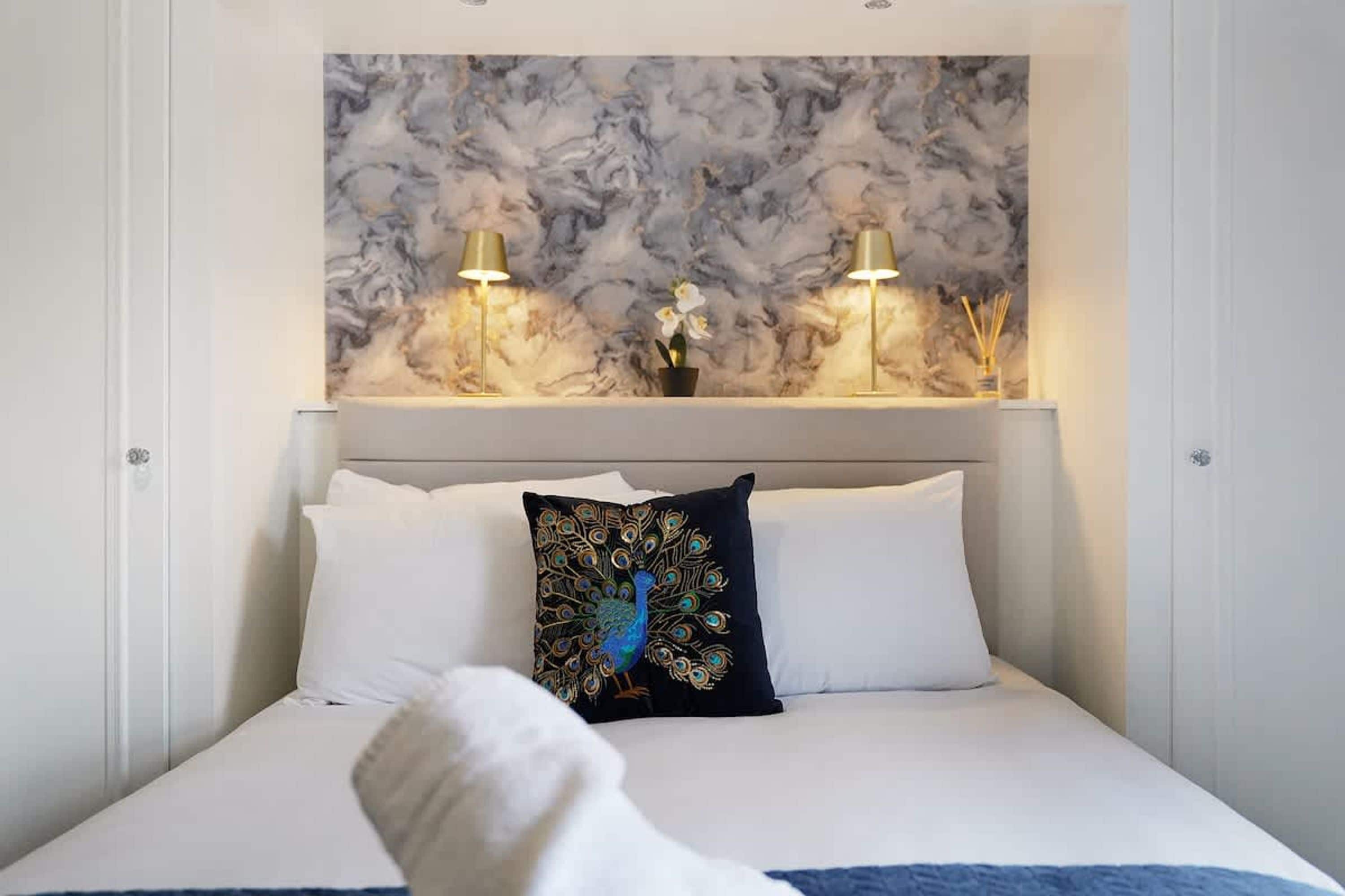 A hotel room with a marble-patterned wall, two bedside lamps, a bed with white bedding, and a decorative pillow with a peacock design.
