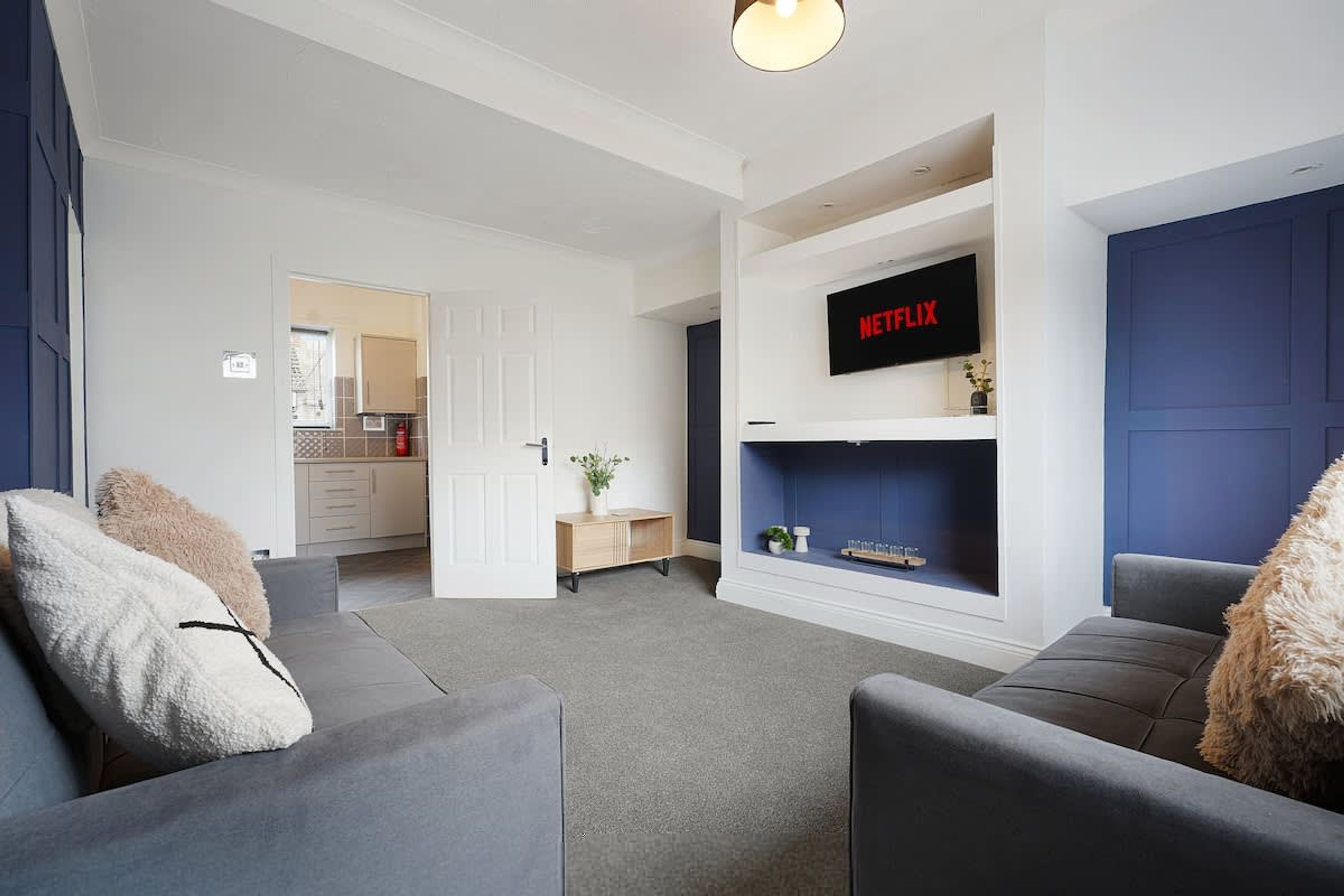 Ein modernes Wohnzimmer mit einem grauen Sofa, einer weißen Tür und einem an der Wand montierten Fernseher, der 'NETFLIX' zeigt.
