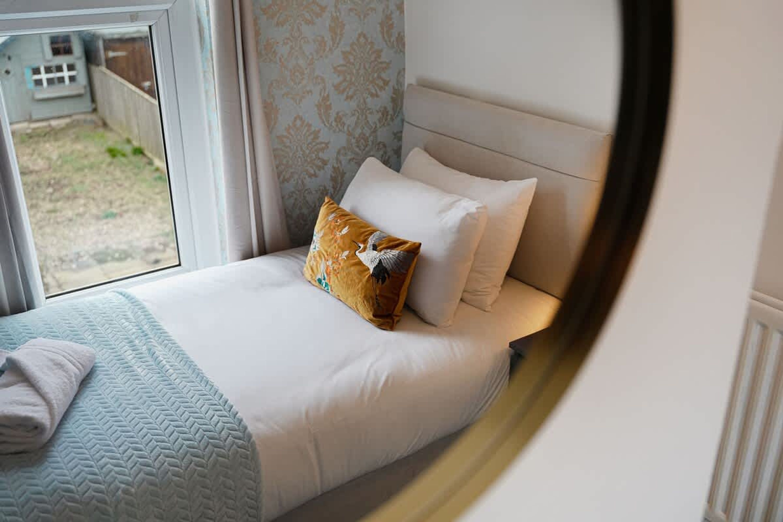 Een bed met wit beddengoed en een kussen met een patroon, een raam met uitzicht op een omheind gebied, en een handdoek op het bed.