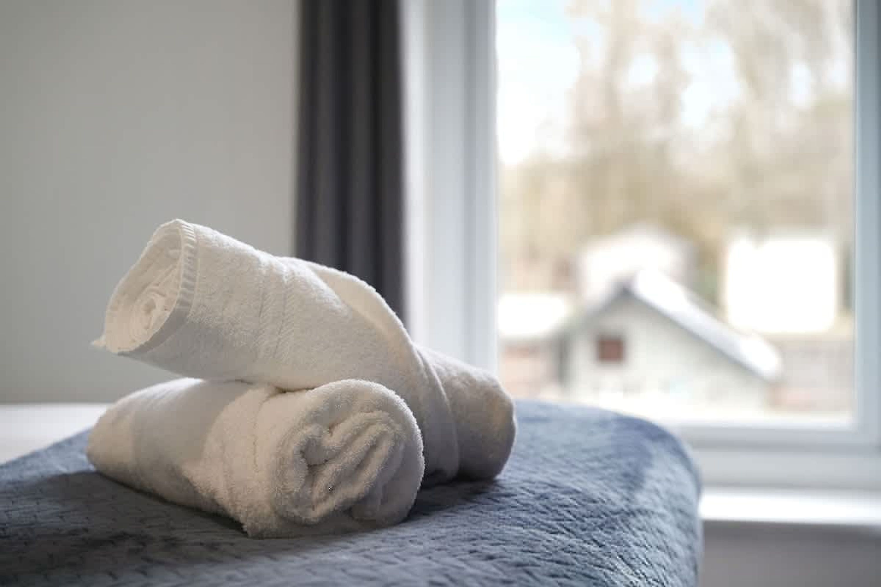 A neatly folded white towel on a textured surface, likely a bed, with a window in the background.
