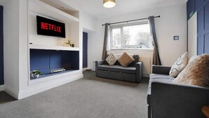 Ein Wohnzimmer mit grauem Sofa, einem flachbildfernseher an einer weißen Wand und einem Fenster mit Vorhängen.