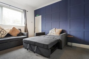 Een moderne hotelkamer met een grijze bank, een bed met grijze beddengoed, een klein rond tafeltje en een blauwe accentmuur.