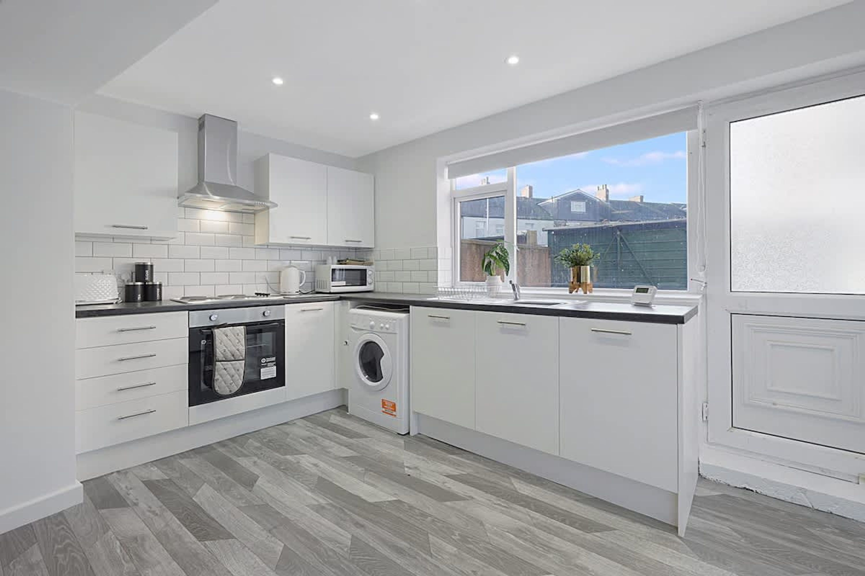 A modern kitchen with white cabinetry, a built-in oven, microwave, and washing machine, and a large window providing a view of the outside.