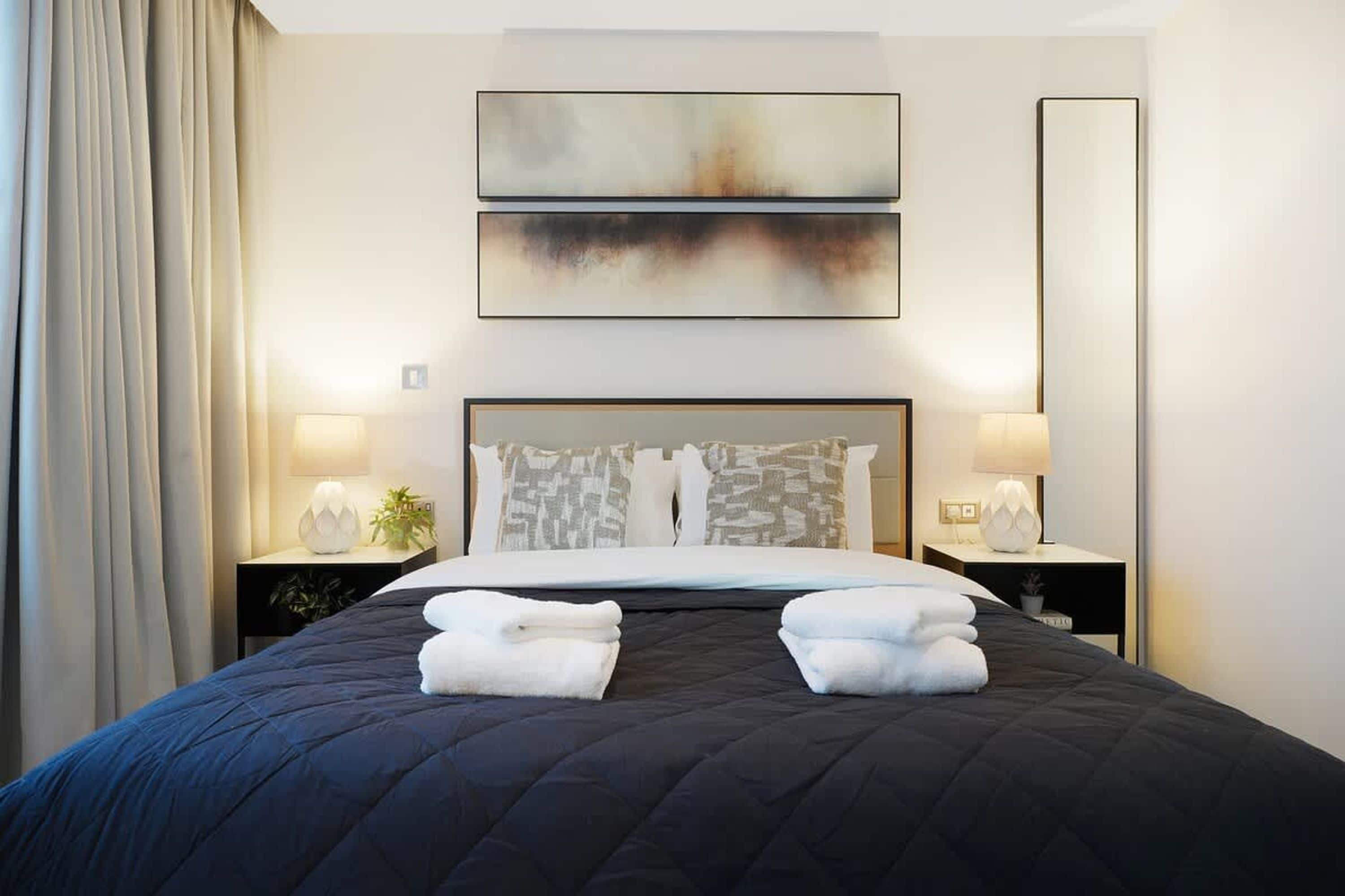 A hotel room with a large bed, two bedside tables with lamps, a painting above the bed, and a view of the cityscape.