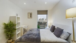 A modern bedroom with a bed, bedside table, lamp, and a view of the city through a window. - Perfect for Couples and Working Away in Manchester (Salford)