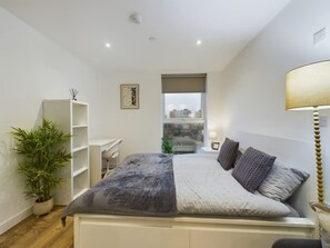 Ein modernes Schlafzimmer mit Bett, Nachttisch, Lampe und Blick auf die Stadt durch ein Fenster.