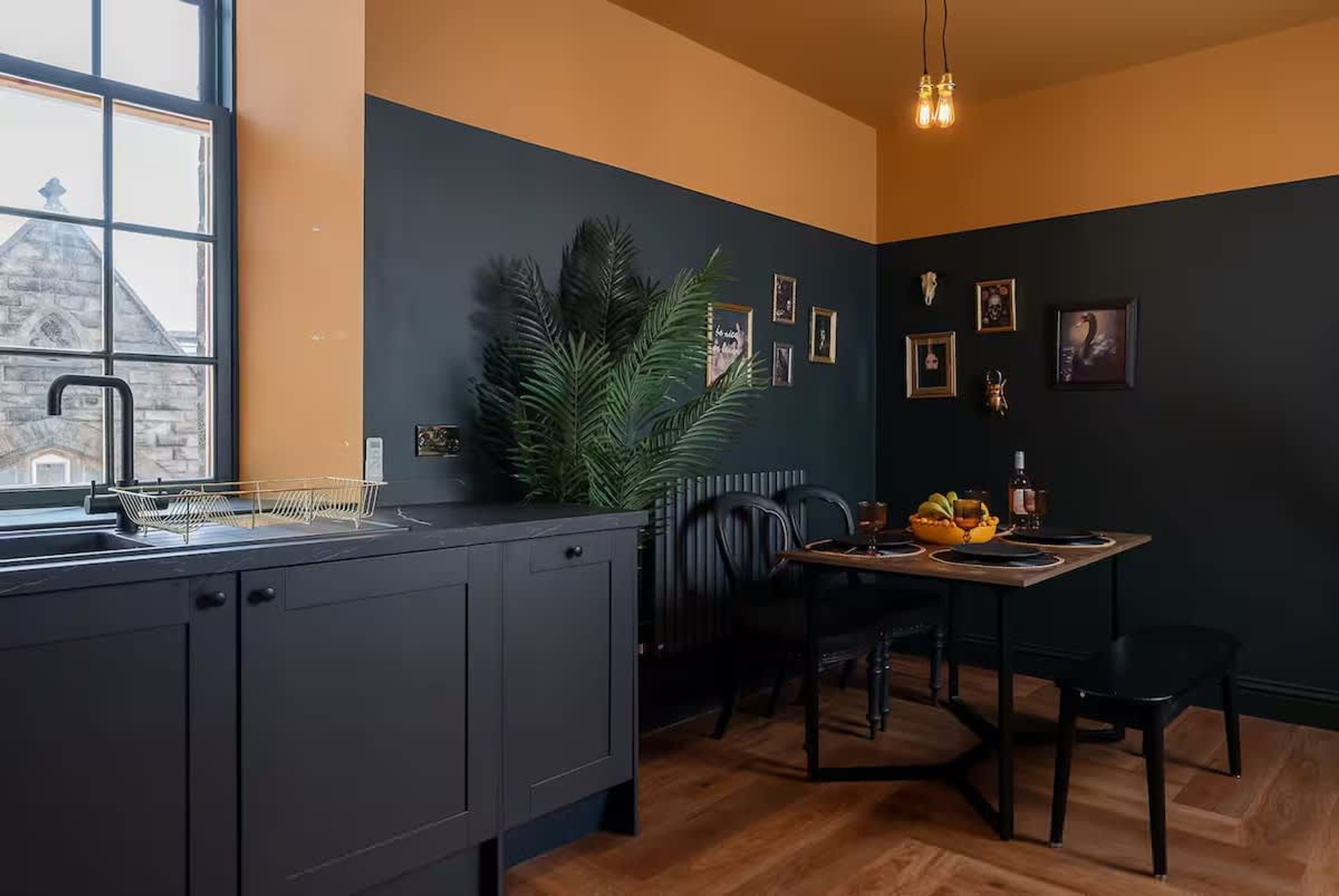 A modern kitchen with dark cabinetry, a dining area with a wooden table and chairs, and a window overlooking a stone building.