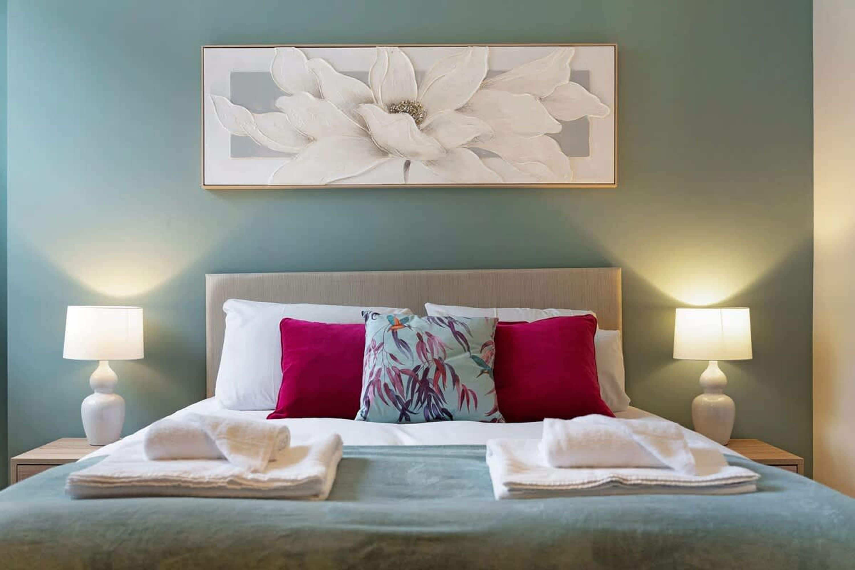 A neatly made bed with pillows and folded towels, a large floral artwork above it, and two bedside lamps on either side.
