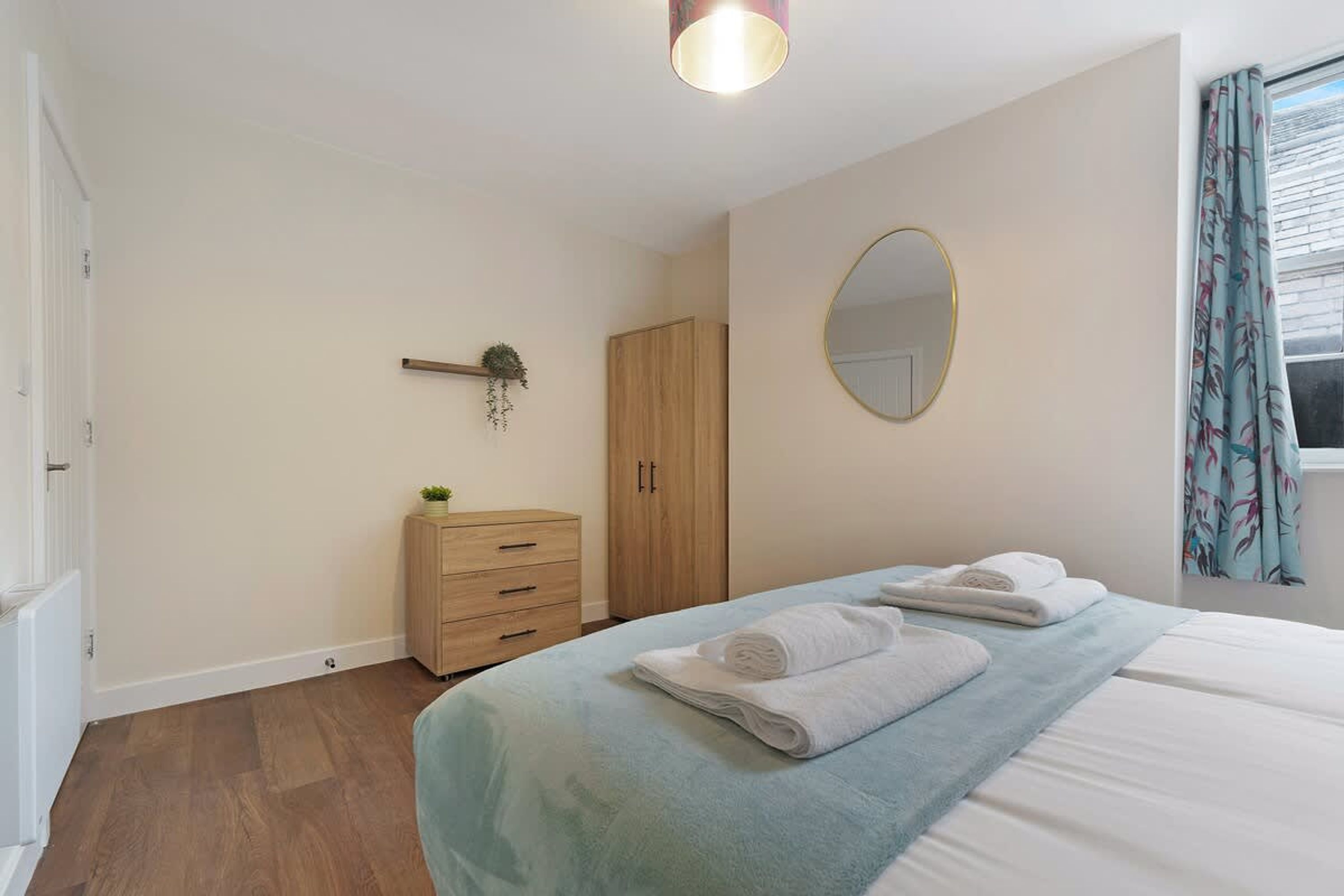 A bedroom with a bed, a dresser, a wardrobe, a mirror, and a window with curtains.
