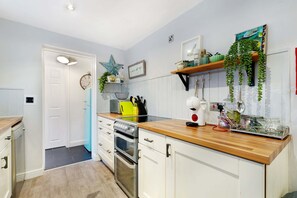 A kitchen with white cabinets, a wooden countertop, a built-in oven, and a microwave. There is a star decoration on the wall, a clock, and a shelf with plants and kitchen utensils. - Coastal 3-bed Home for Families Contractors (Redcar)